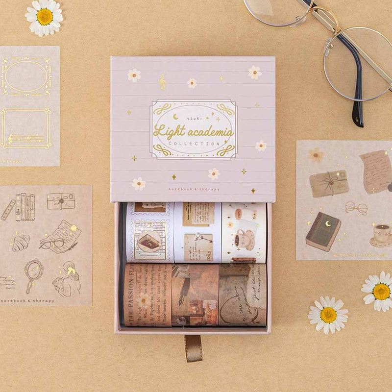 Tsuki Light Academia Washi Tape Set by Notebook Therapy on brown background with a pair of glasses and some pressed daisies around the open box
