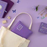 Notebook pouch in a canvas tote bag on purple background with enchanted garden washi tapes, stamps and notebook in the background