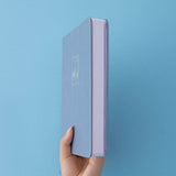 A hand holding a blue leather notebook showing the white page edges