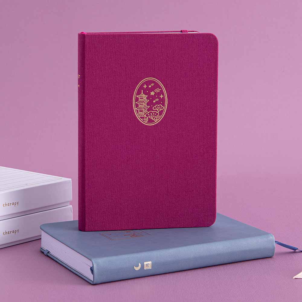 Tsuki Kyoto bullet journal with a pink linen cover and gold details standing on a blue Tokyo notebook