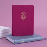 Tsuki Kyoto bullet journal with a pink linen cover and gold details standing on a blue Tokyo notebook