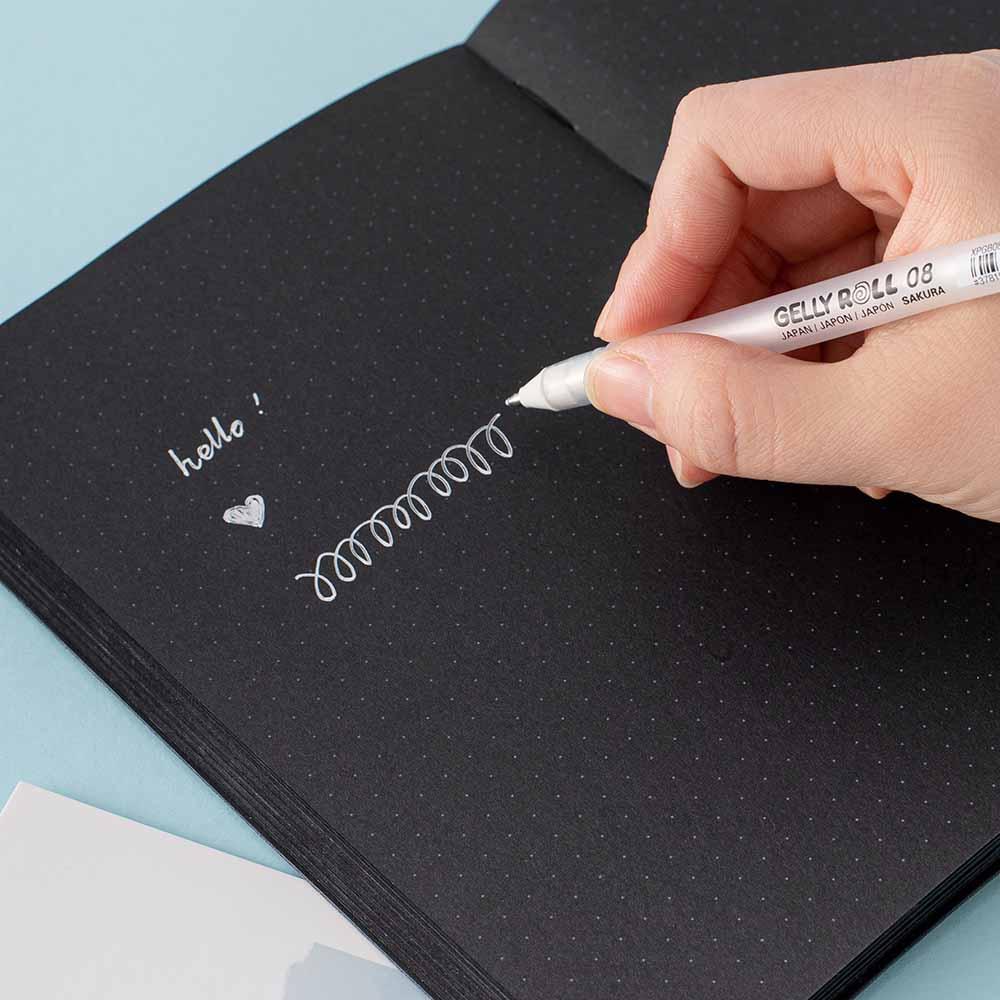 Close up of white Gelly Roll Pen held in hand open Tsuki Black Paper Limited Edition Hardcover Bullet Journal on light teal background