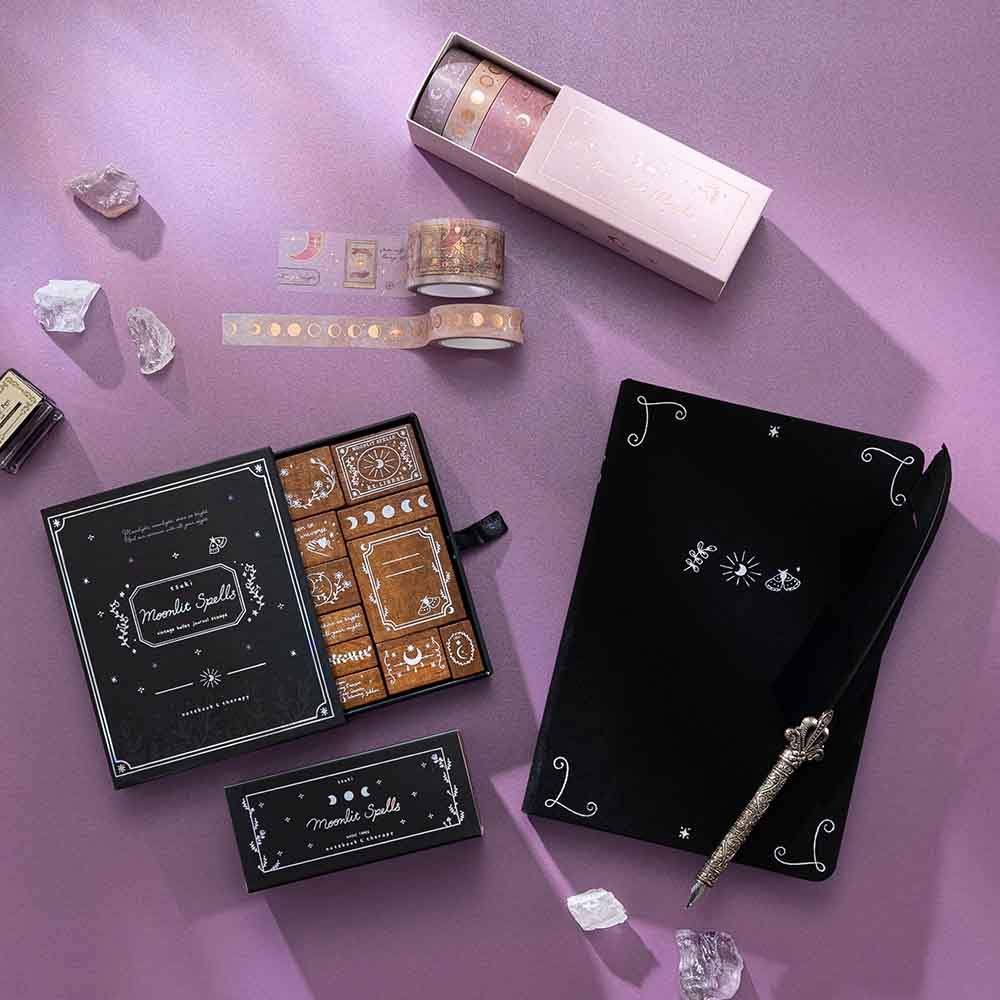 Tsuki ‘Moonlit Spells’ Bullet Journal Stamp Set with Tsuki ‘Moonlit Spell’ Limited Edition Holographic Bullet Journal and Tsuki ‘Moonlit Blush’ Washi Tape Set and Tsuki ‘Moonlit Spells’ Washi Tape Set with amethyst stones and quill pen on purple background