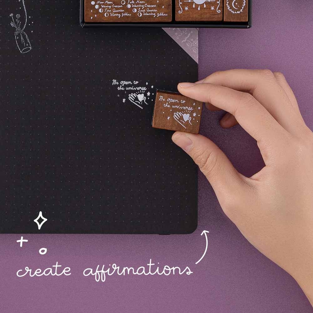 Close up of Tsuki ‘Moonlit Spells’ Bullet Journal Stamps to create affirmations with held in hand on open black page bullet journal on purple background