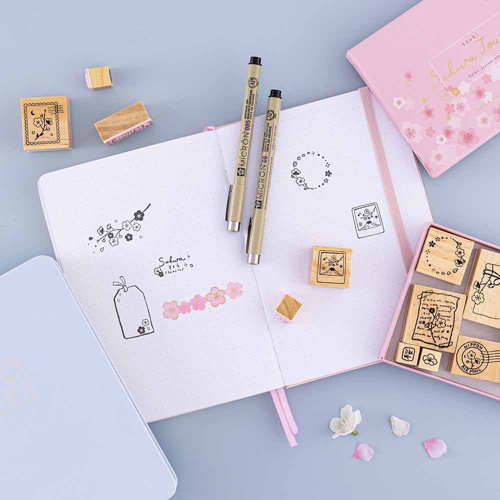 Tsuki ‘Sakura Journey’ Bullet Journal Stamp Set on open Tsuki ‘Lunar Blossom’ Limited Edition Bullet Journal with Tsuki ‘Sakura Journey’ Limited Edition Bullet Journal with cherry blossom petals and two pens on light blue background