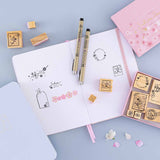 Tsuki ‘Sakura Journey’ Bullet Journal Stamp Set on open Tsuki ‘Lunar Blossom’ Limited Edition Bullet Journal with Tsuki ‘Sakura Journey’ Limited Edition Bullet Journal with cherry blossom petals and two pens on light blue background