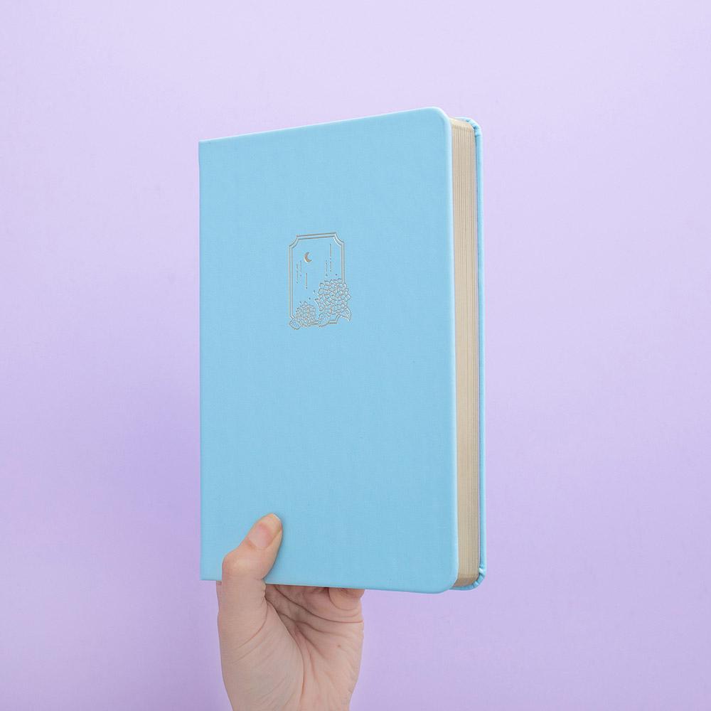 Tsuki Endless Summer Limited Edition Bullet Journal in Petal Blue held in hands at spine angle in lilac background
