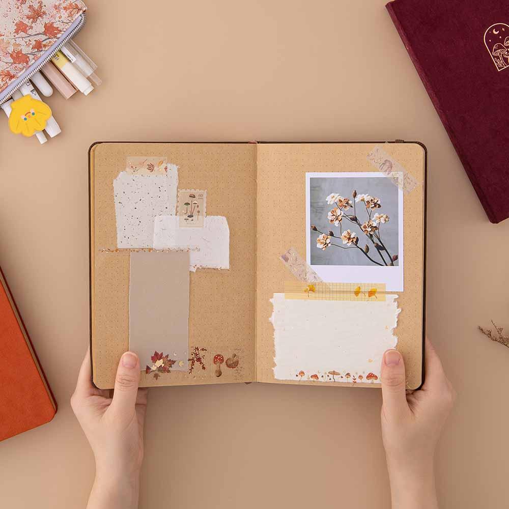 Open Tsuki Kraft Paper Limited Edition Bullet Journal with Tsuki Mixed Scrapbook Paper and Tsuki Handmade Petal Paper and polaroid picture and Tsuki ‘Maple Dreams’ Washi Tapes on page held in hands with Tsuki ‘Maple Dreams’ Pop-Up Pencil Case in maple and Tsuki ‘Kinoko’ Limited Edition Bullet Journal and Tsuki ‘Kitsune’ Limited Edition Fox Bullet Journal on beige background