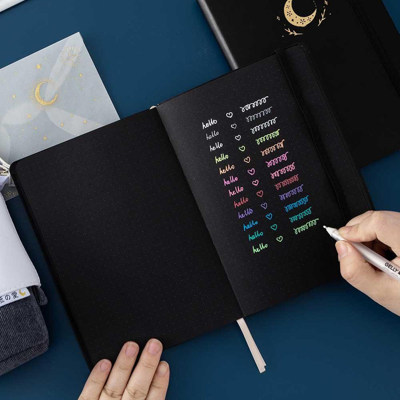Open Tsuki Black Paper Limited Edition Hardcover Bullet Journal with Gelly Roll Pen held in hands with Tsuki Pop-Up Pencil case in charcoal on dark blue background