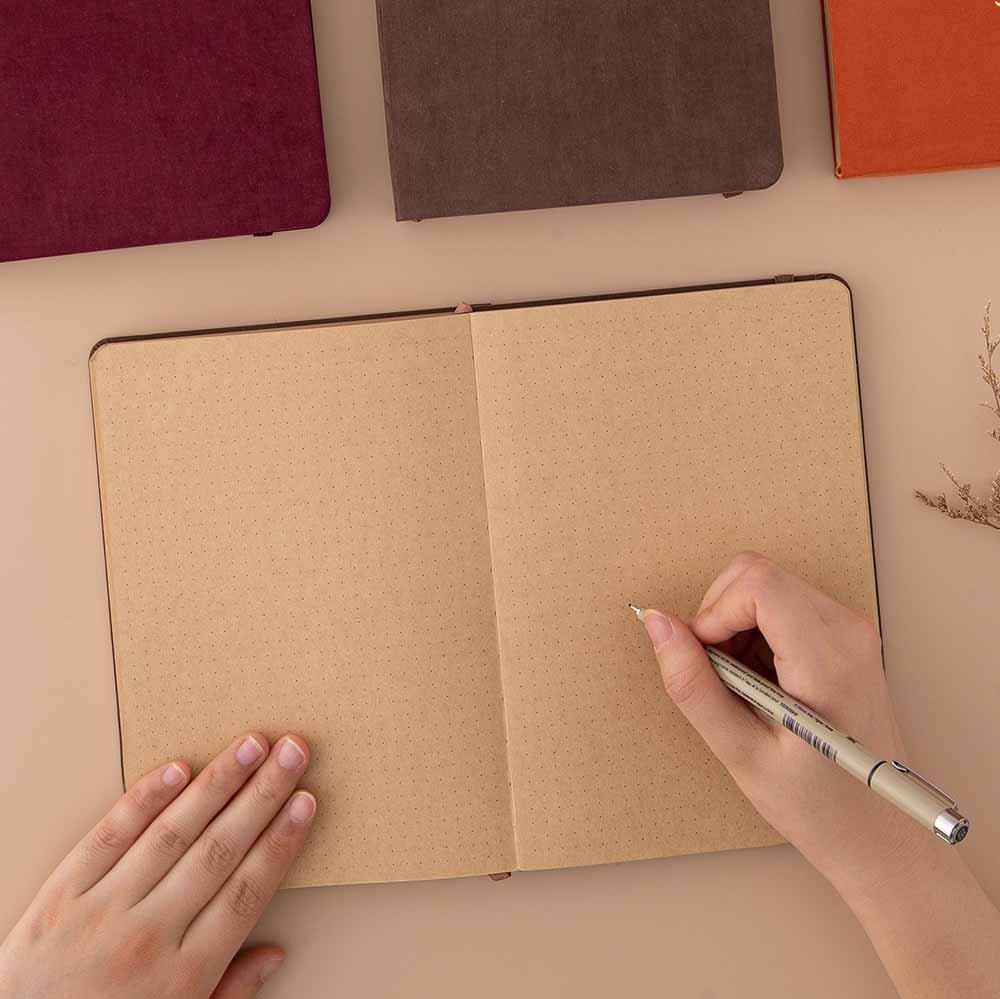 Open Tsuki Kraft Paper Limited Edition Bullet Journal with pen held in hands with Tsuki ‘Kitsune’ Limited Edition Fox Bullet Journal and Tsuki ‘Nara’ Limited Edition Bullet Journal and Tsuki ‘Kinoko’ Limited Edition Bullet Journal on beige background