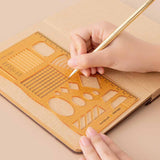 Tsuki Bullet Journal Stencil Set in neutral demonstrated on open bullet journal kraft paper page spread with pencil held in hands on beige background