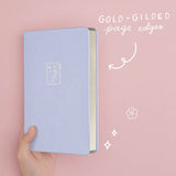 Tsuki ‘Sakura Journey’ Limited Edition Bullet Journal held in hand at spine angle with gold gilded page edges in pink background