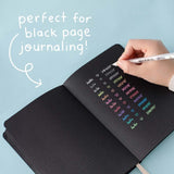 Gelly Roll Pen in white held in hand over open Tsuki Black Paper Limited Edition Hardcover Bullet Journal on light teal background