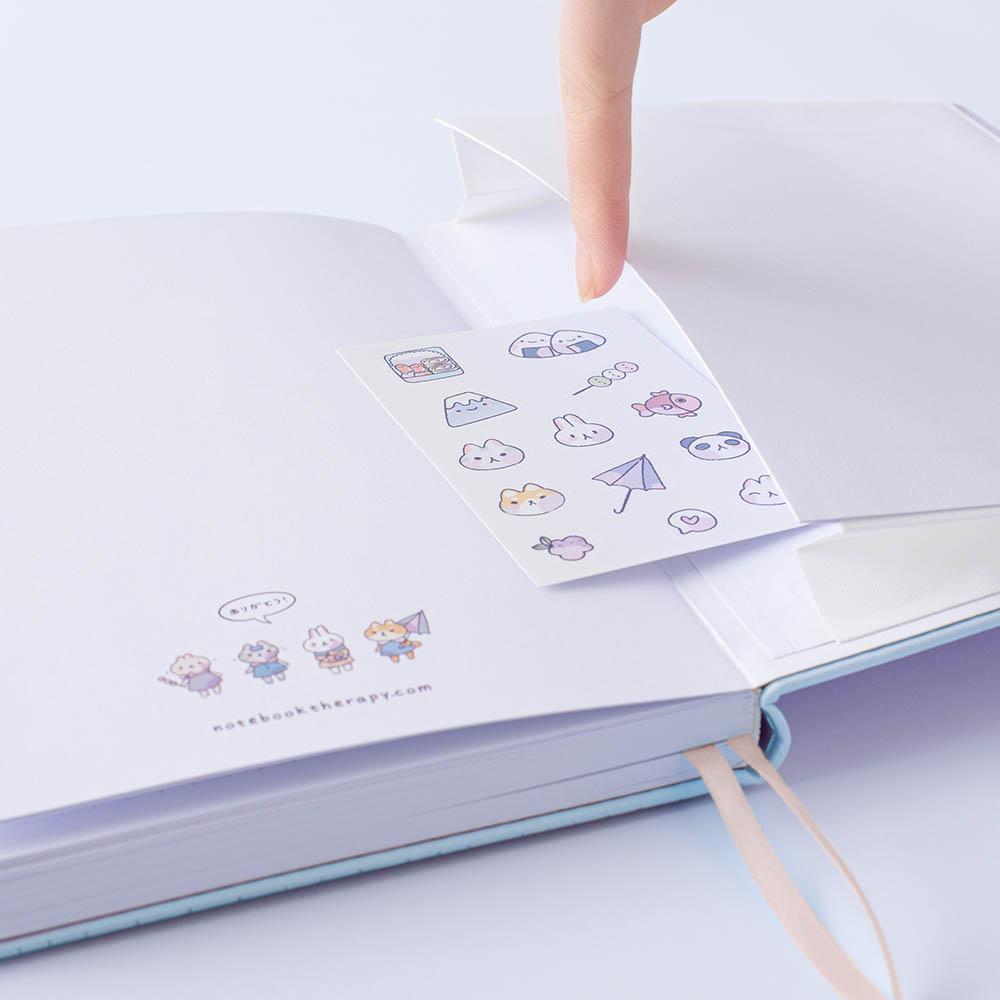 Close up of open back page pocket of Tsuki ‘Four Seasons: Autumn Edition’ Bullet Journal made with milkkoyo held in hand with free stickers sheet on lilac background
