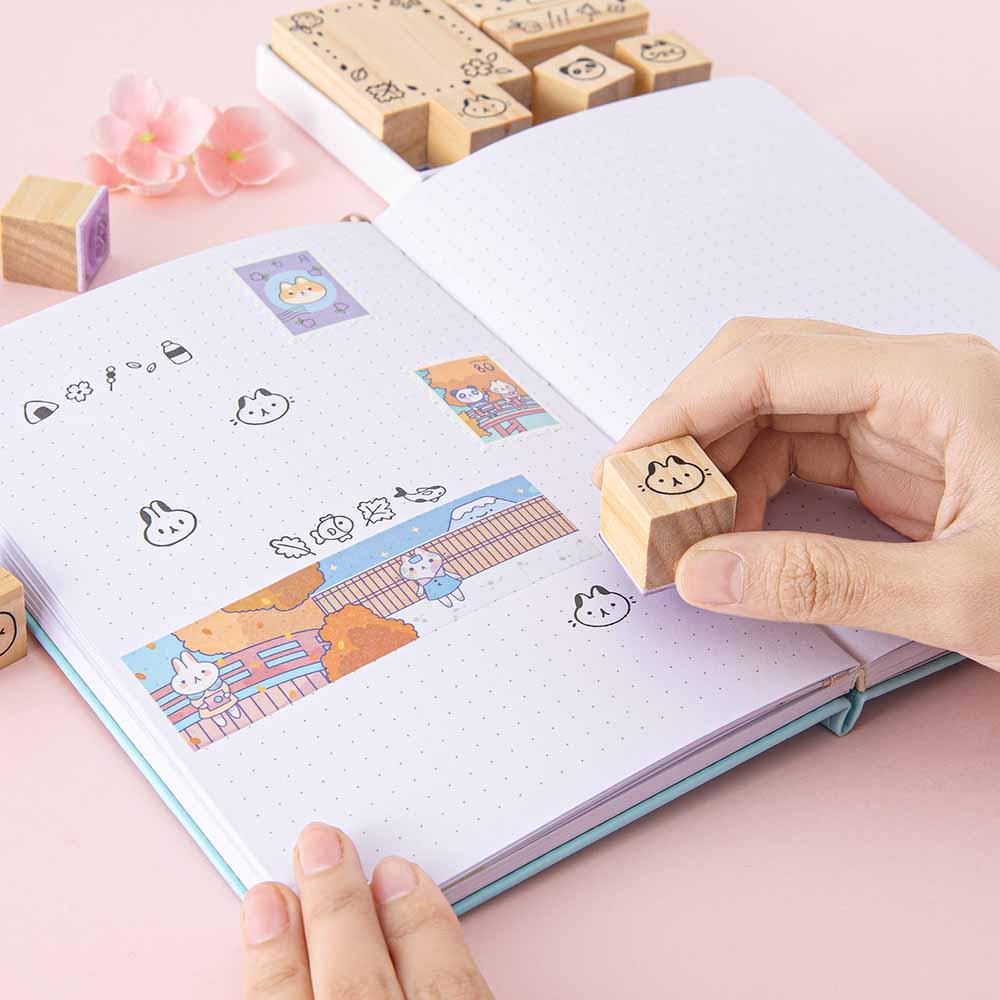 Tsuki ‘Four Seasons’ Bullet Journal Stamp Set by Notebook Therapy x Milkkoyo held in hand with Tsuki ‘Four Seasons’ Washi Tape on open bullet journal spread with pink flower on petal pink background