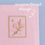 Close up of Tsuki ‘Sakura Journey’ Notebook Pouch with embroidered stamp design on light blue background