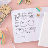 Close up of Tsuki ‘Flutter + Dream’ Bullet Journal Stamp Set by Notebook Therapy x Pelinkan with frame stamps on open Tsuki Teal Sky ‘Flutter + Dream’ Limited Edition Bullet Journal by Notebook Therapy x Pelinkan with Tsuki Cloud White White Sky ‘Flutter + Dream’ Limited Edition Bullet Journal by Notebook Therapy x Pelinkan with gelly pen on pastel pink background