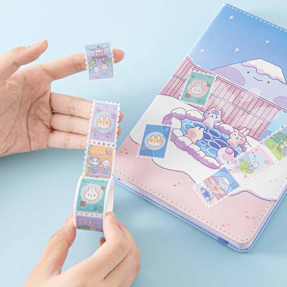 Tsuki ‘Four Seasons’ Stamp Washi Tape by Notebook Therapy x Milkkoyo held in hands with Tsuki ‘Four Seasons: Winter Edition’ Bullet Journal on light blue background