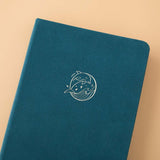 Close up of the front cover of Sea green velvet Tsuki Dolphin Days bullet journal on peach background