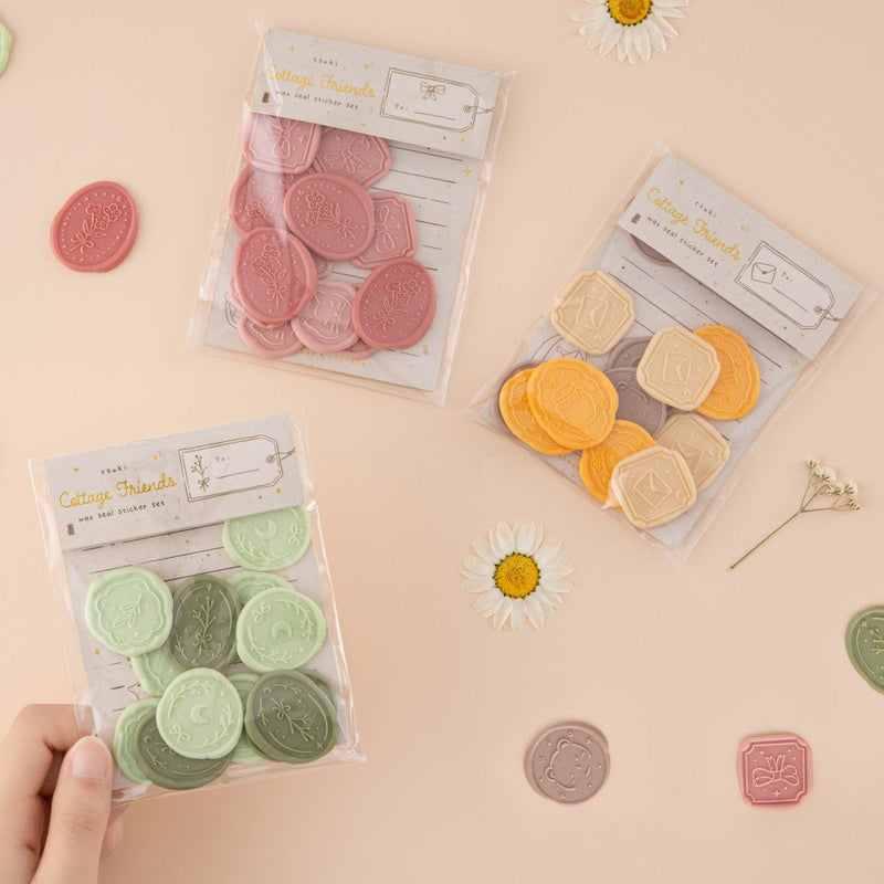 Hand holding green wax seal sticker set with pink wax seall sticker set and cream, yellow and brown in the background