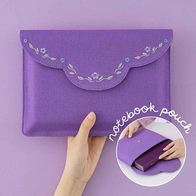 Hand holding a purple fabric notebook envelope pouch with flower embroidery and an image in the middle of a notebook fitting in the envelope notebook pouch