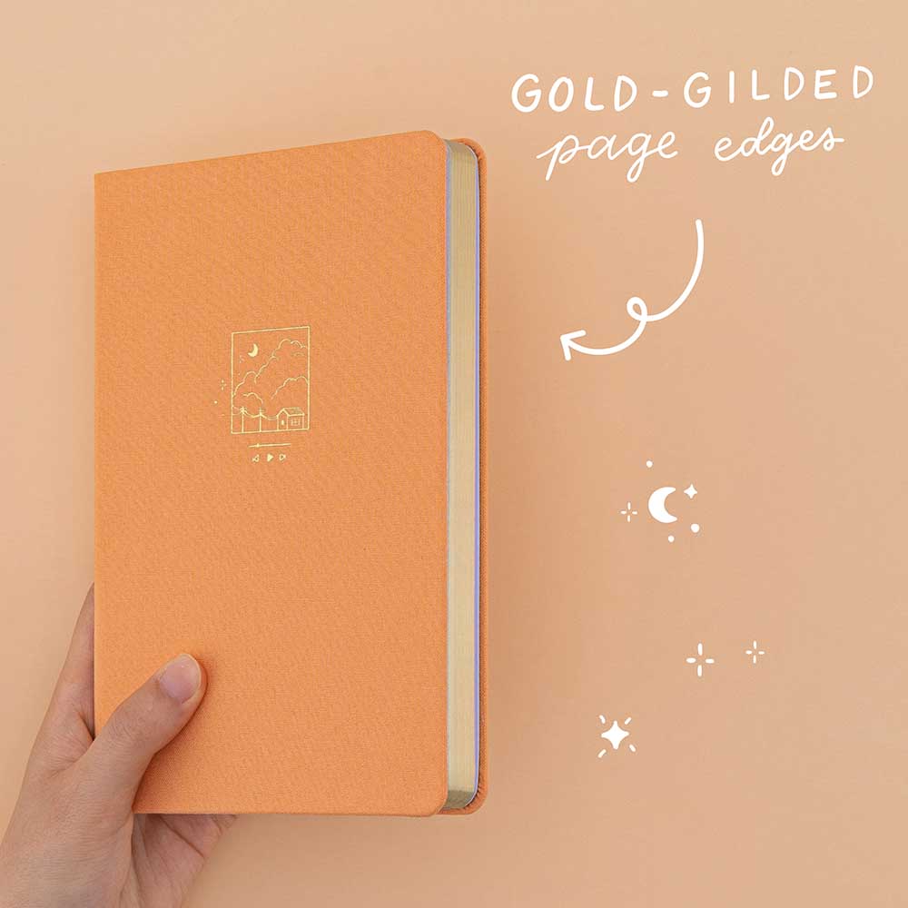 Hand holding Tsuki Golden Hour bullet journal at an angle showing the gold gilded page edges 