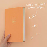 Hand holding Tsuki Golden Hour bullet journal at an angle showing the gold gilded page edges