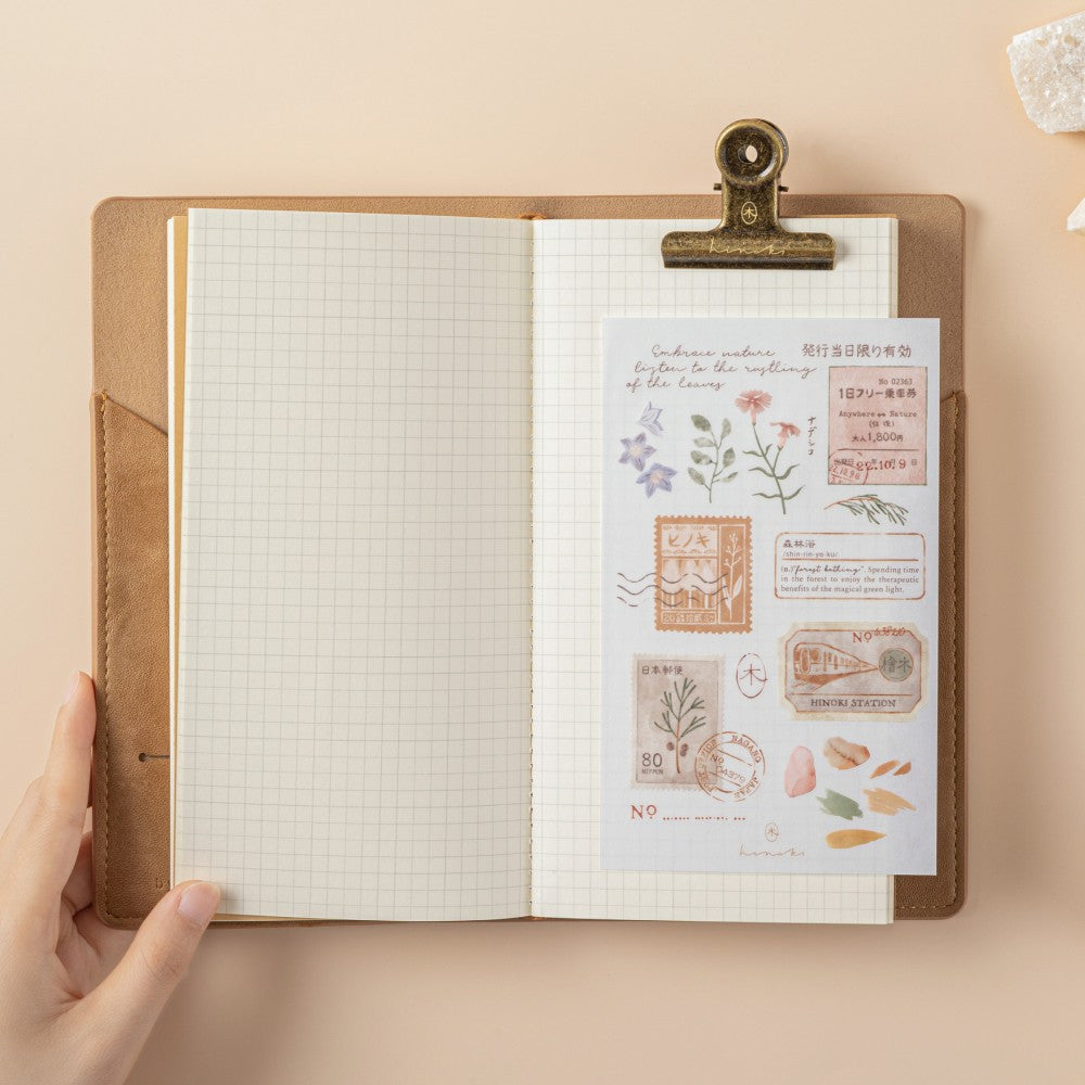 Open Travel Notebook with a brown leather case and a blank paper insert, a sticker tucked in the pocket and pine leaves tucked in the cardholder