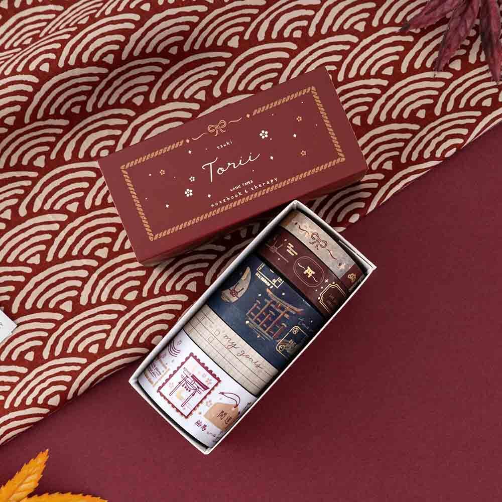 An opened box of Tsuki Torii washi tape set by notebook therapy laid out on red background with red traditional Japanese patterned fabric