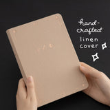 Hands holding Stardust Dawn Bullet Journal with white text that reads “hand-crafted linen cover”