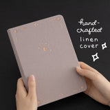 Hands holding Tsuki Moonlit Dusk Bullet Journal with white text that reads “hand-crafted linen cover”