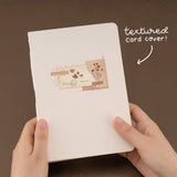 Hands holding the Junk Journal notebook with white text “textured card cover”