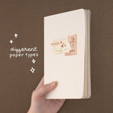 Hand holding up the Junk Journal notebook against brown background with lettering “different paper types”