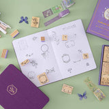 Tsuki ‘Enchanted Garden’ Stamp Set on dotted notebook and sage green background with purple flower decoration, stamps and washi tapes 