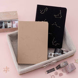 Tsuki Constellations Collection by Notebook Therapy including 2x bullet journals, 1 in beige 1 in black and a washi tape set in a basket on pink background