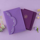 Purple envelope pouch with embroidered details on top of 2 purple notebooks with lavender gold foil design