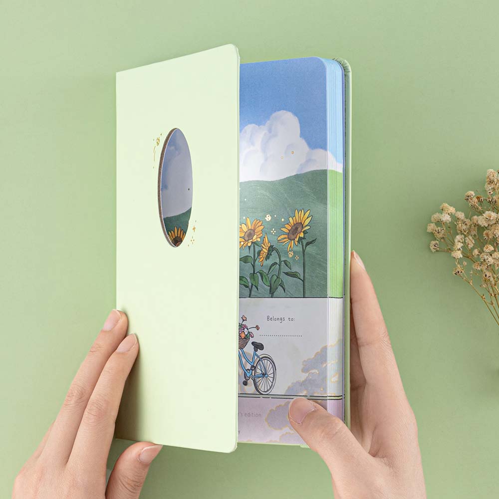 Hands holding Tsuki Four Seasons Summer Collectors Edition 2022 sage bullet journal notebook with a cutout in the middle and sunflower illustrations on the first cover