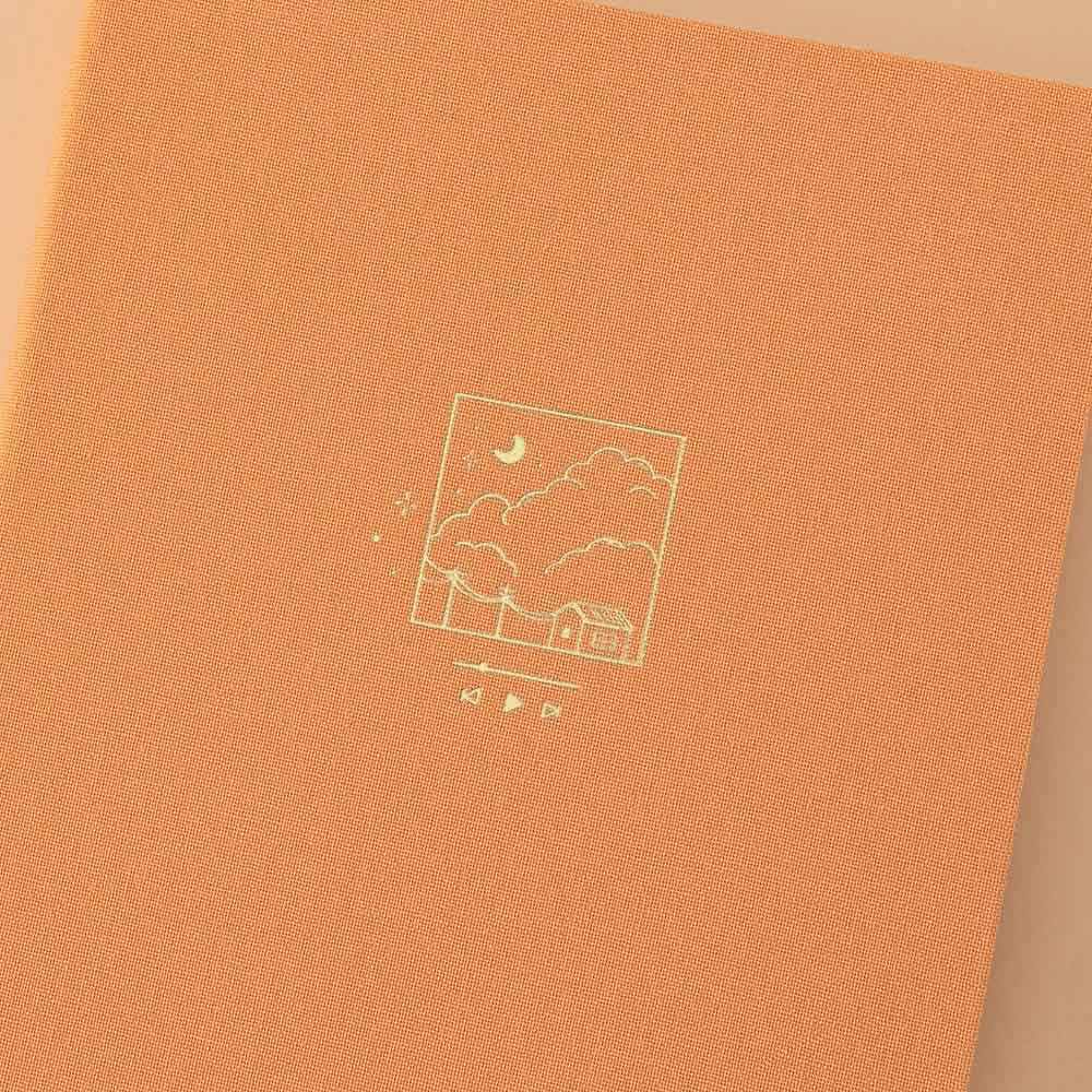 Close up of Tsuki Golden Hour bullet journal design which is an illustration of a house and some clouds in a box frame with a playlist bar underneath in gold foil on orange linen
