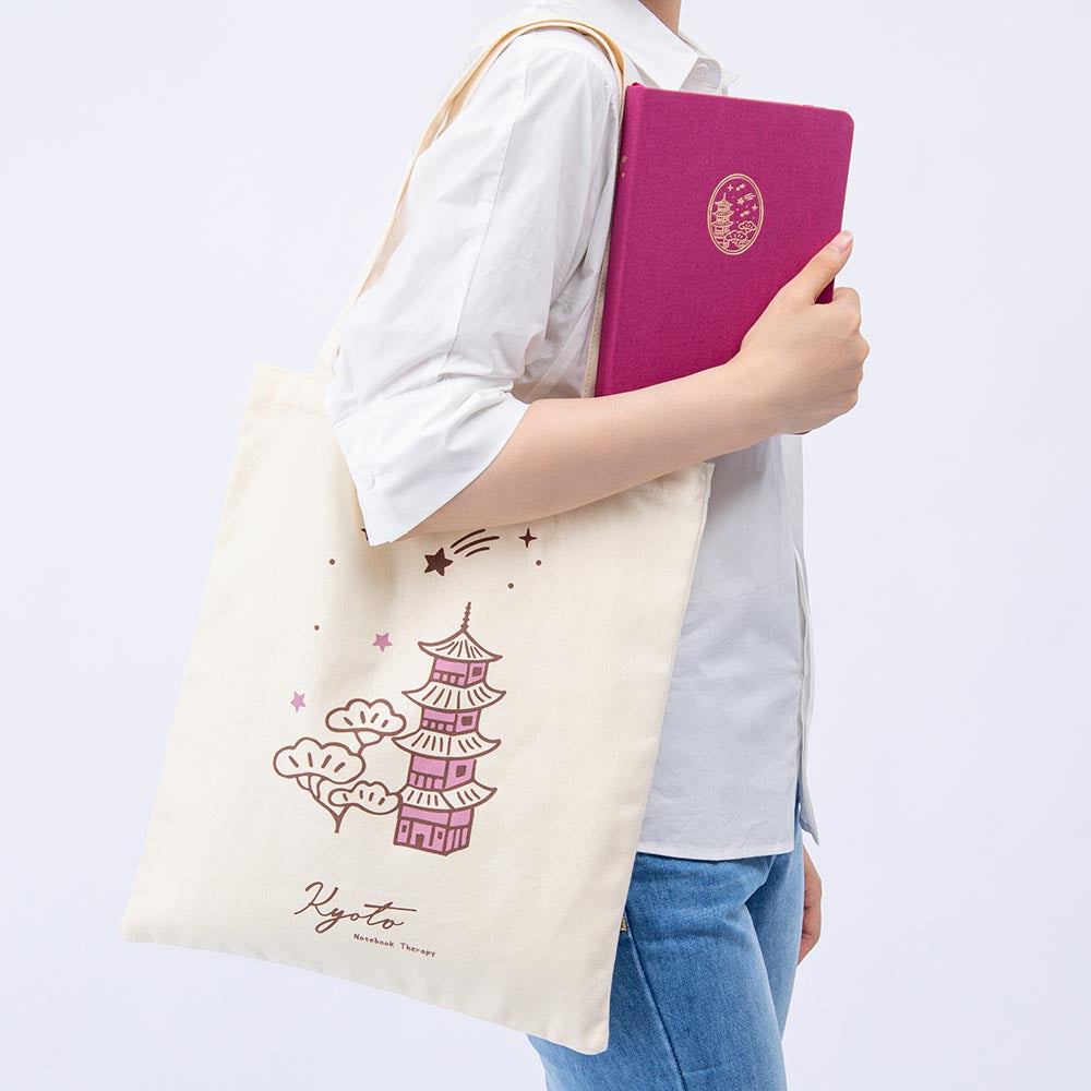 Woman carrying a fuchsia pink notebook and a canvas tote bag with a temple design and the word “Kyoto” written on it
