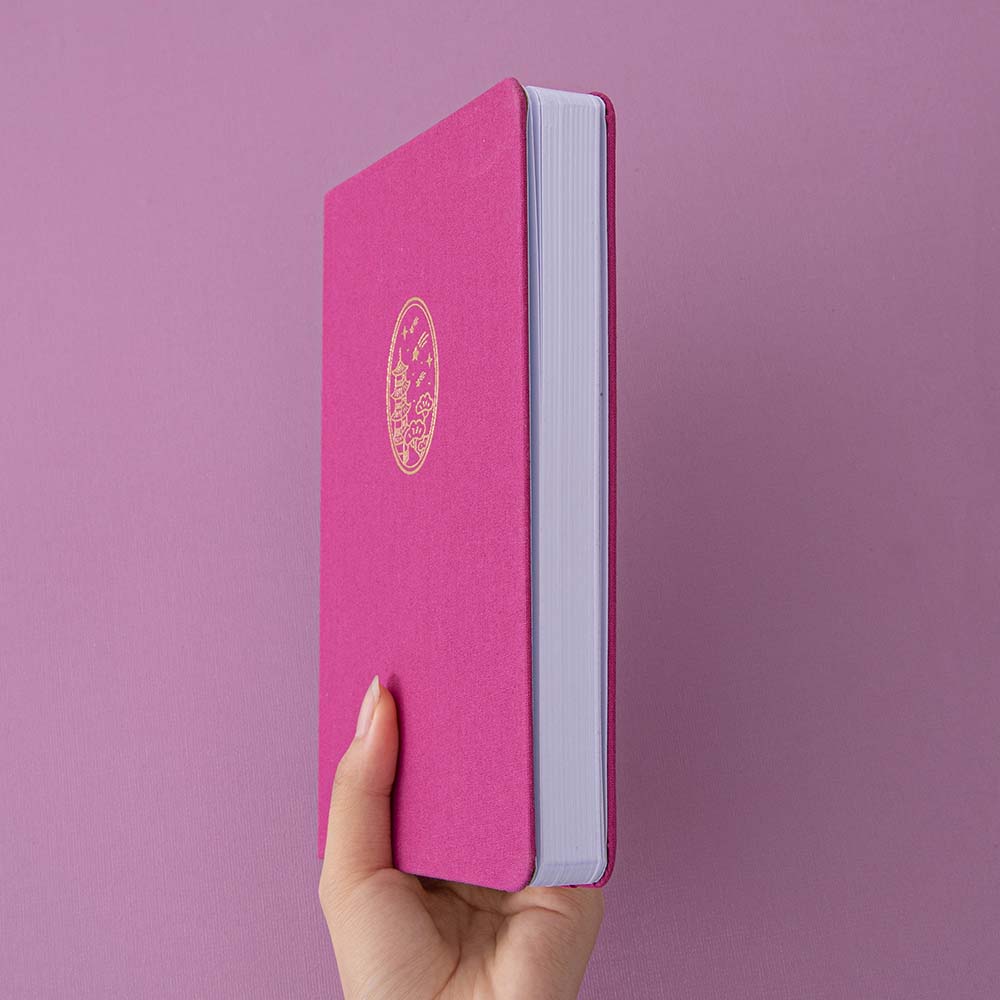 A hand holding a fuchsia pink bullet journal notebook against a pink background showing the white page edges