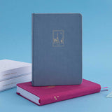 Tsuki Tokyo bullet journal with a blue faux leather cover standing on a pink Kyoto notebook