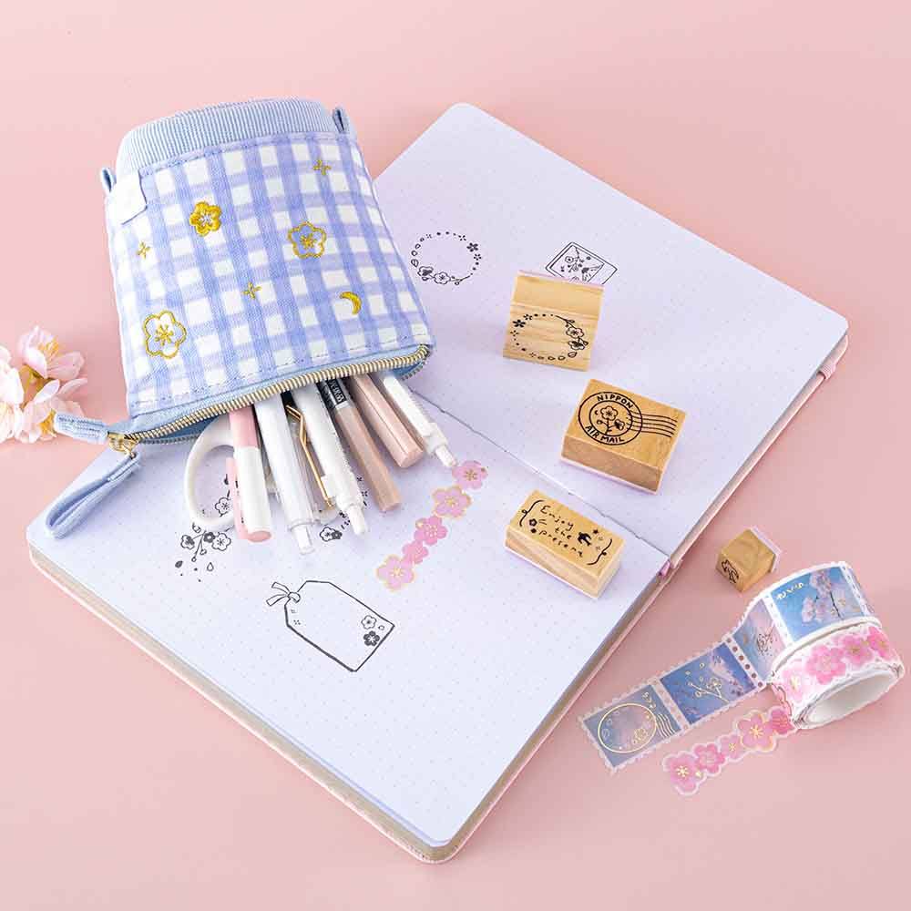 Tsuki ‘Sakura Journey’ Pop-Up Pencil Case with pens inside on open Tsuki ‘Lunar Blossom’ Limited Edition Bullet Journal with Tsuki ‘Sakura Journey’ Washi Tapes and Tsuki ‘Sakura Journey’ Bullet Journal Stamps with cherry blossoms on light pink background