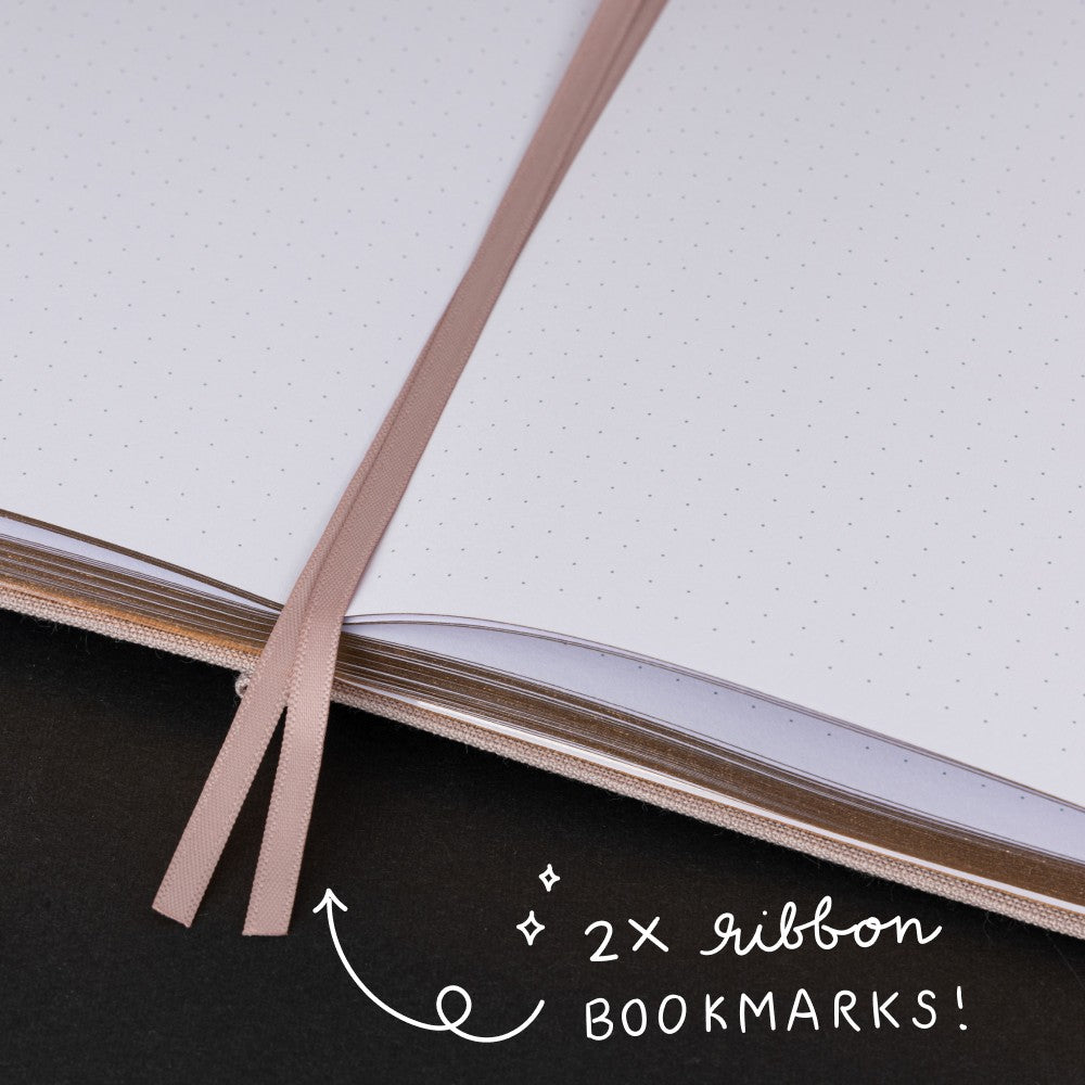 Feather quill and rose gold moon-shaped paperclip on white page dotted notebook with lettering “luxury 160gsm pages”
