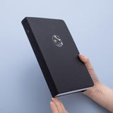 Tsuki deep black Playful Orca limited edition notebook held in hands at an angle in blue background