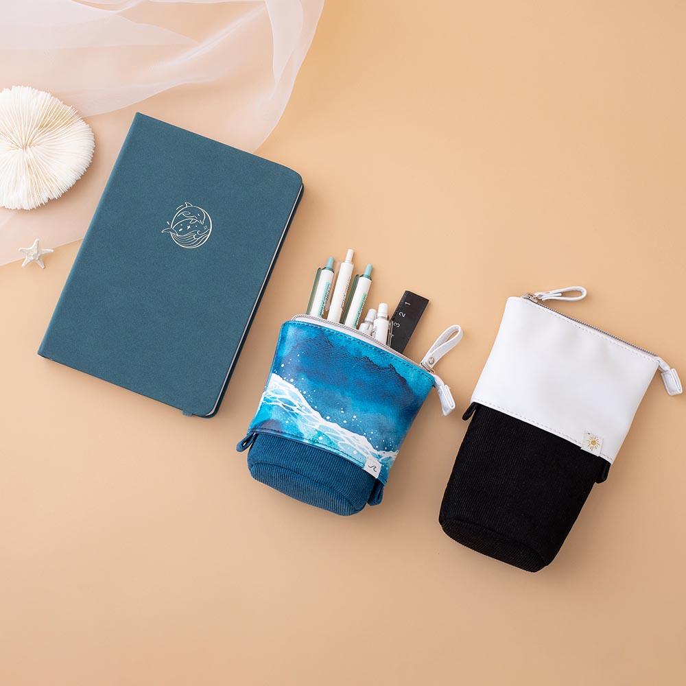 Tsuki Ocean Edition pop up standing pencil cases in Orca Black and Ocean Blue at an angle with sea green textured vegan leather Dolphin Days notebook with pens and seashell on peach background