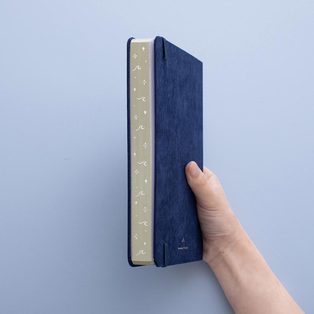 Tsuki deep blue textured vegan leather Gentle Giant luxury edition notebook with gold page edges held in hand at an angle in blue background