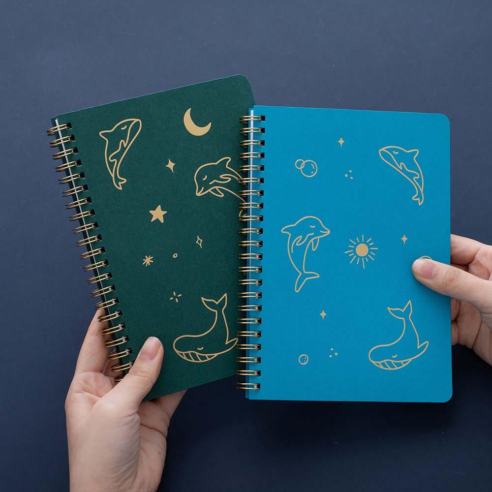 Tsuki Ocean Edition Ring Bound notebooks in aqua blue and deep teal held in hands in dark blue background