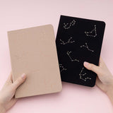 A pair of hands holding 2 tsuki Constellations bullet journal, 1 in beige and 1 in black on pink background