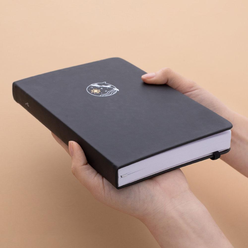 Tsuki deep black Playful Orca limited edition notebook held in hands at an angle in peach background