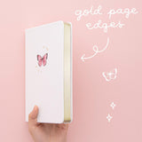 Tsuki Cloud White ‘Flutter + Dream’ Limited Edition Bullet Journal by Notebook Therapy x Pelinkan with gold gilded page edges held in hand in pastel pink background